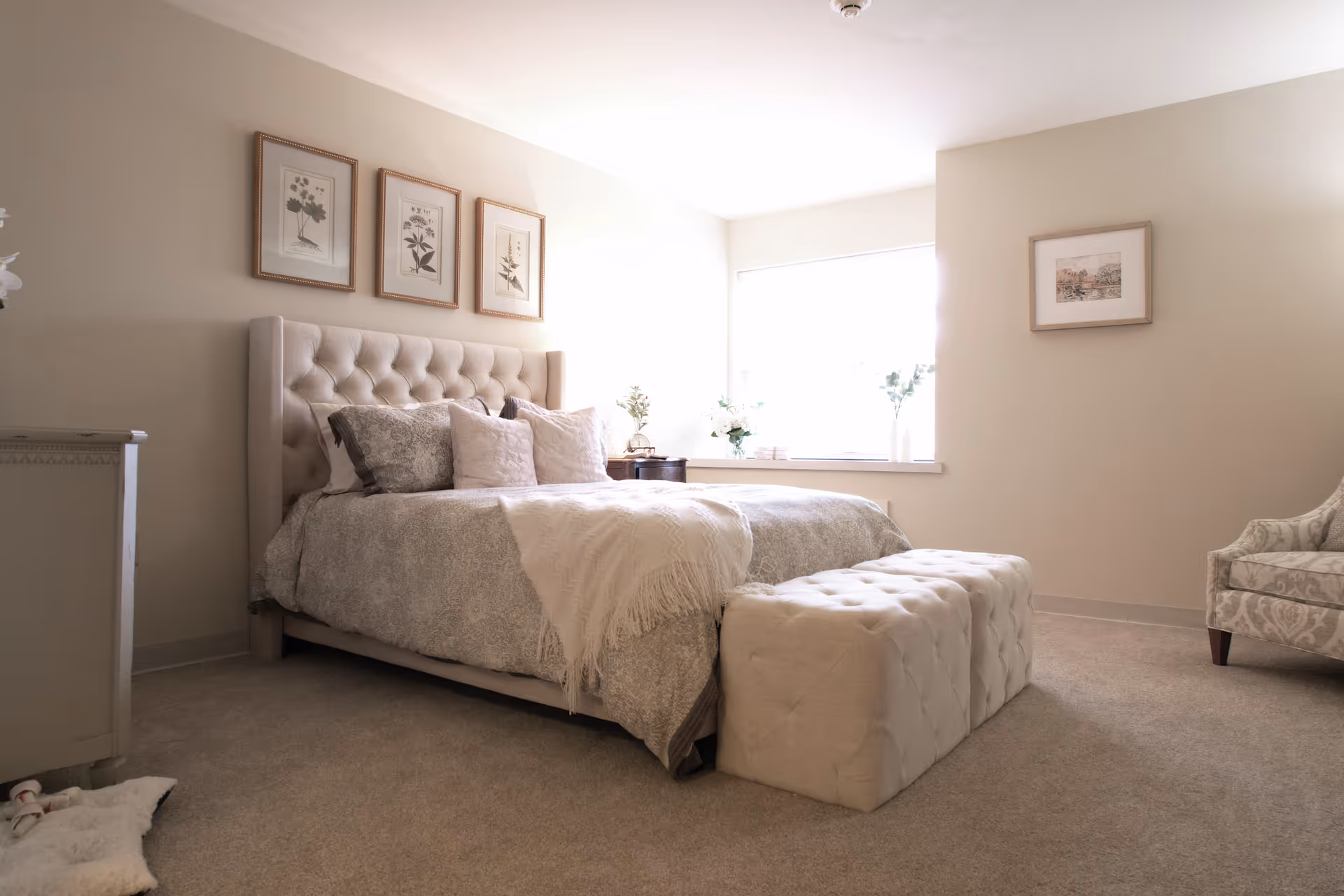 A cozy bedroom with a large tufted bed adorned with multiple pillows and a throw blanket. There is a matching tufted bench at the foot of the bed, a cushioned armchair to the right, and framed botanical artwork on the walls. A window lets in natural light, brightening the neutral-toned room.