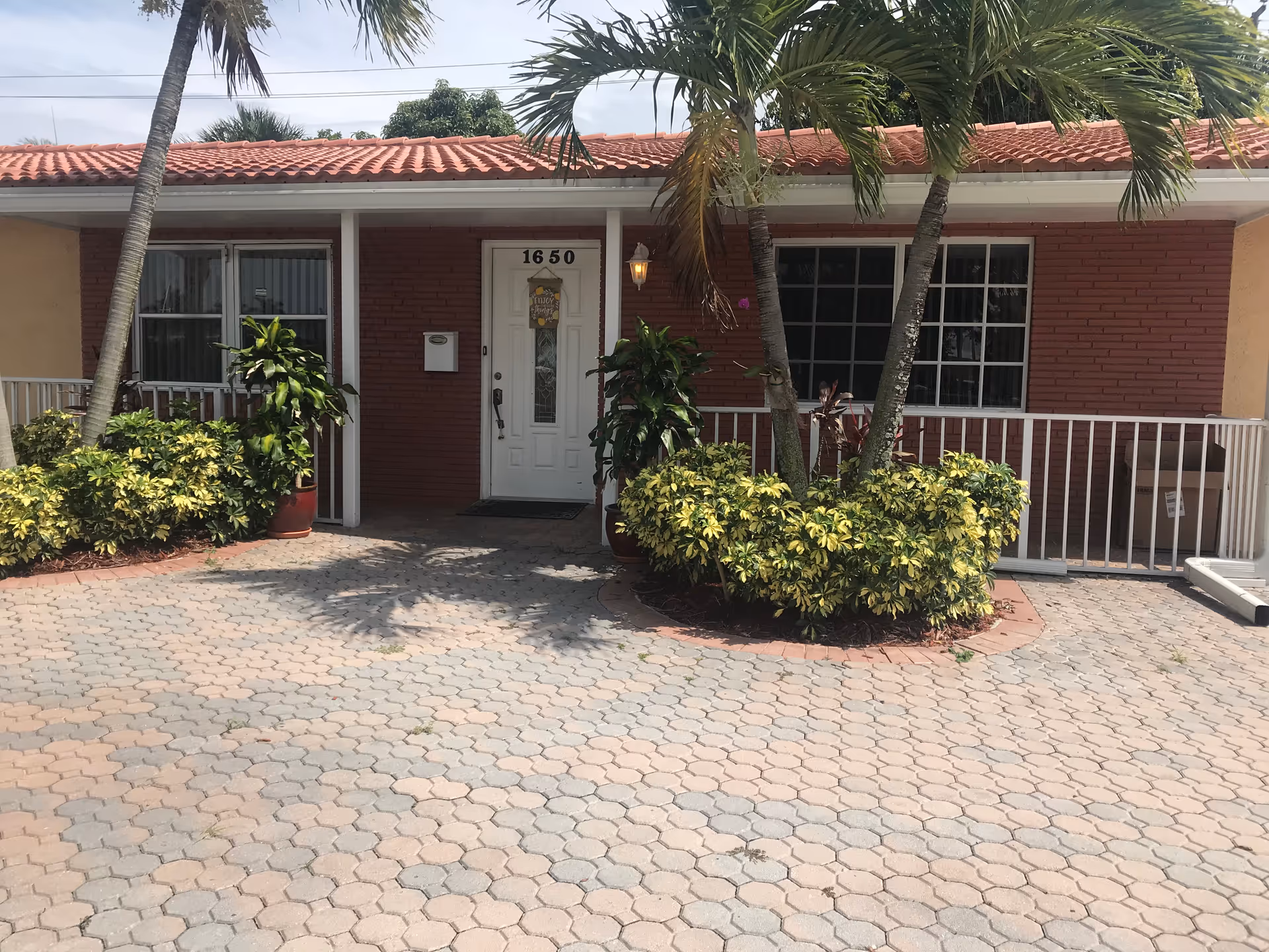 Front exterior view of a single-story building with a red tiled roof and brick walls. The entrance door is white with the number 1650 above it. There are two palm trees and various green plants and shrubs in front of the building, along with a paved driveway or walkway.