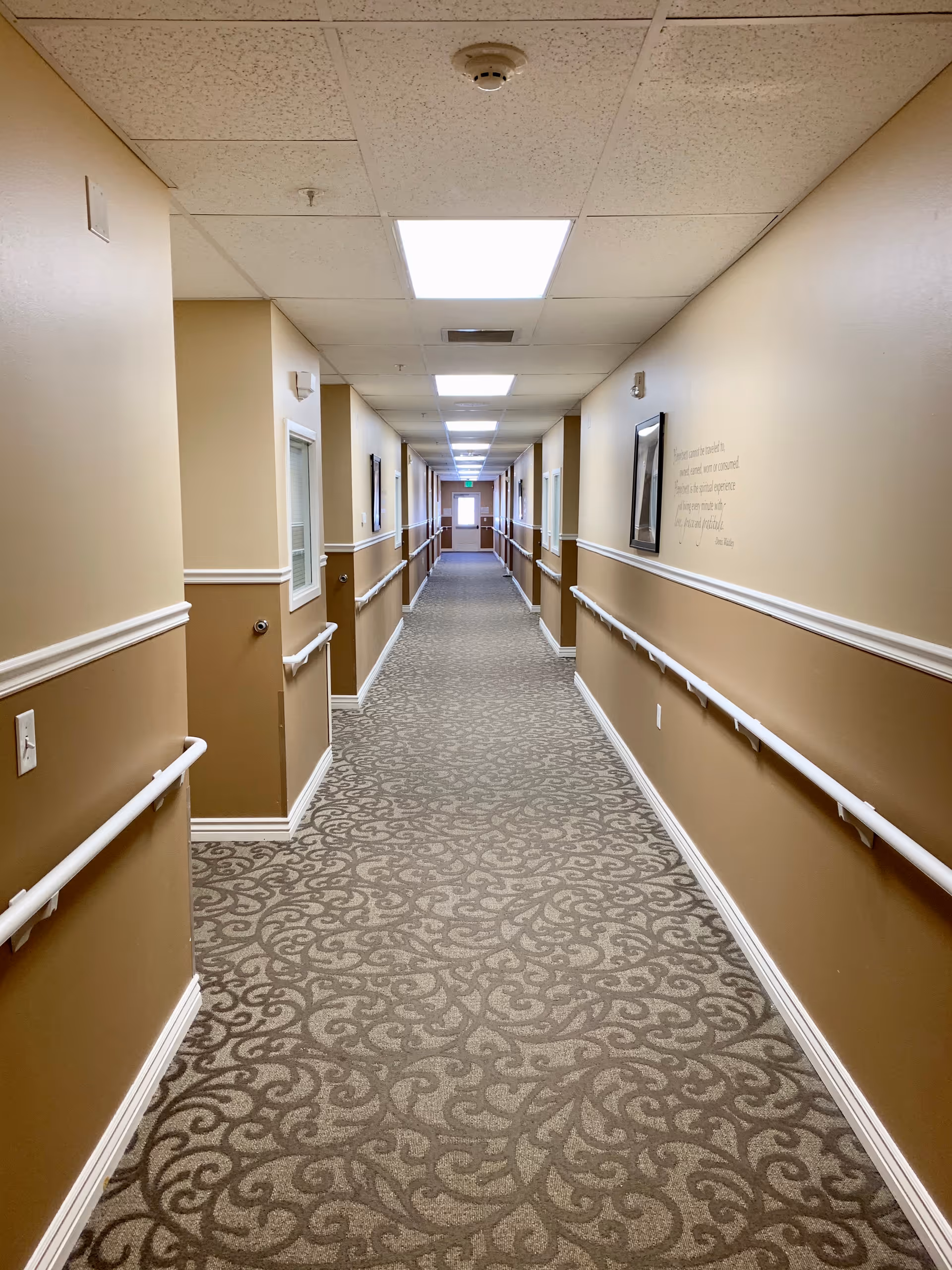 A long, well-lit hallway in an assisted living facility with beige walls, patterned carpet, handrails on both sides, and several doors and framed pictures along the walls. The ceiling has recessed lighting panels.