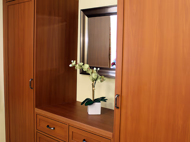 A wooden dresser with drawers and two tall cabinets on either side, featuring a small white vase with green and white flowers on top. Above the dresser is a large rectangular mirror with a metallic frame reflecting part of a window with floral curtains.