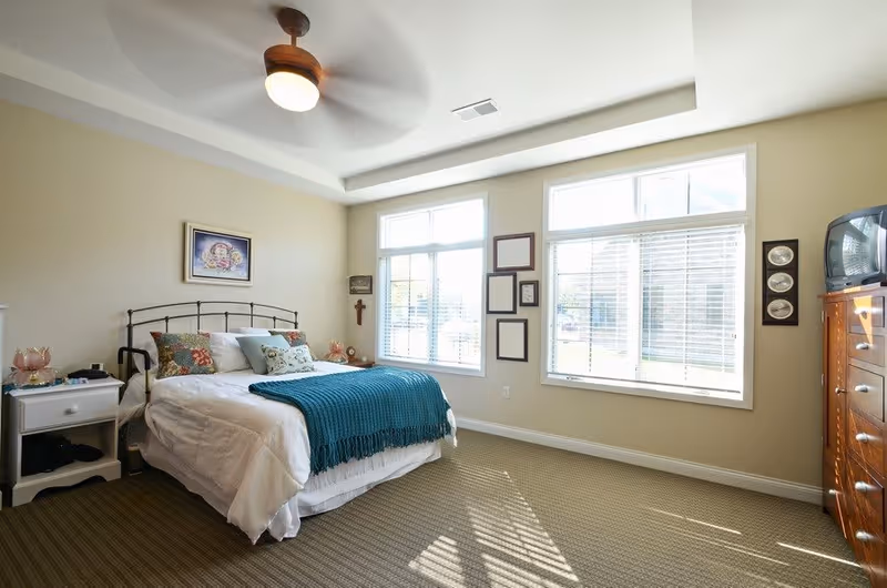 A bright bedroom with a large bed covered in white bedding and a teal throw blanket. The bed has multiple decorative pillows and a black metal headboard. There is a white nightstand with a lamp and other items on the left side of the bed. The room has beige walls, two large windows with white blinds letting in natural light, a ceiling fan with a light, and a wooden dresser with a TV on top on the right side. Several framed pictures and a cross hang on the walls.