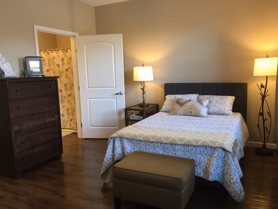 Neatly furnished bedroom with a bed, nightstands and lamps, a dresser and an ottoman, with an open door showing a bathroom shower curtain.