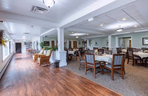 A spacious dining area in a senior living facility with multiple round tables covered with white tablecloths and surrounded by wooden chairs with cushions. To the left, there is a wide hallway with wooden flooring, several armchairs, and large windows letting in natural light. The ceiling has recessed lighting and decorative fixtures, and the walls are painted in a soft green color with white trim.