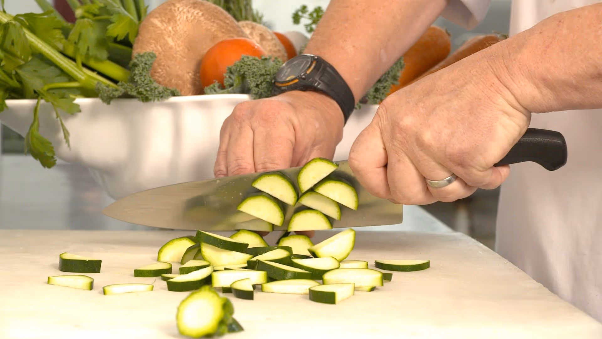 Close-up of hands slicing zucchini on a cutting board with fresh vegetables in the background.