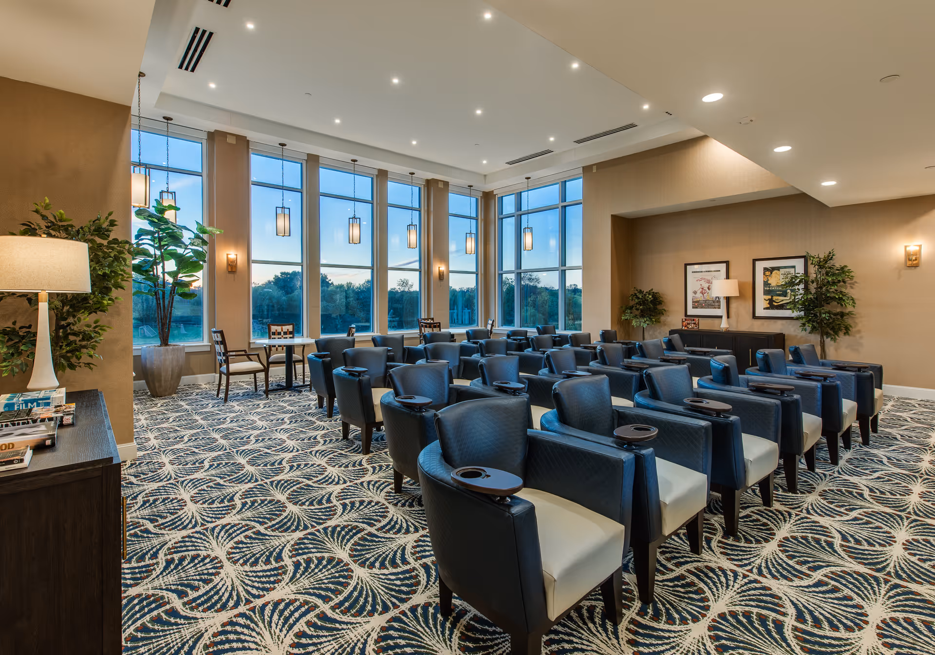 A spacious senior living community meeting room with rows of upholstered armchairs, patterned carpet, and large floor-to-ceiling windows.