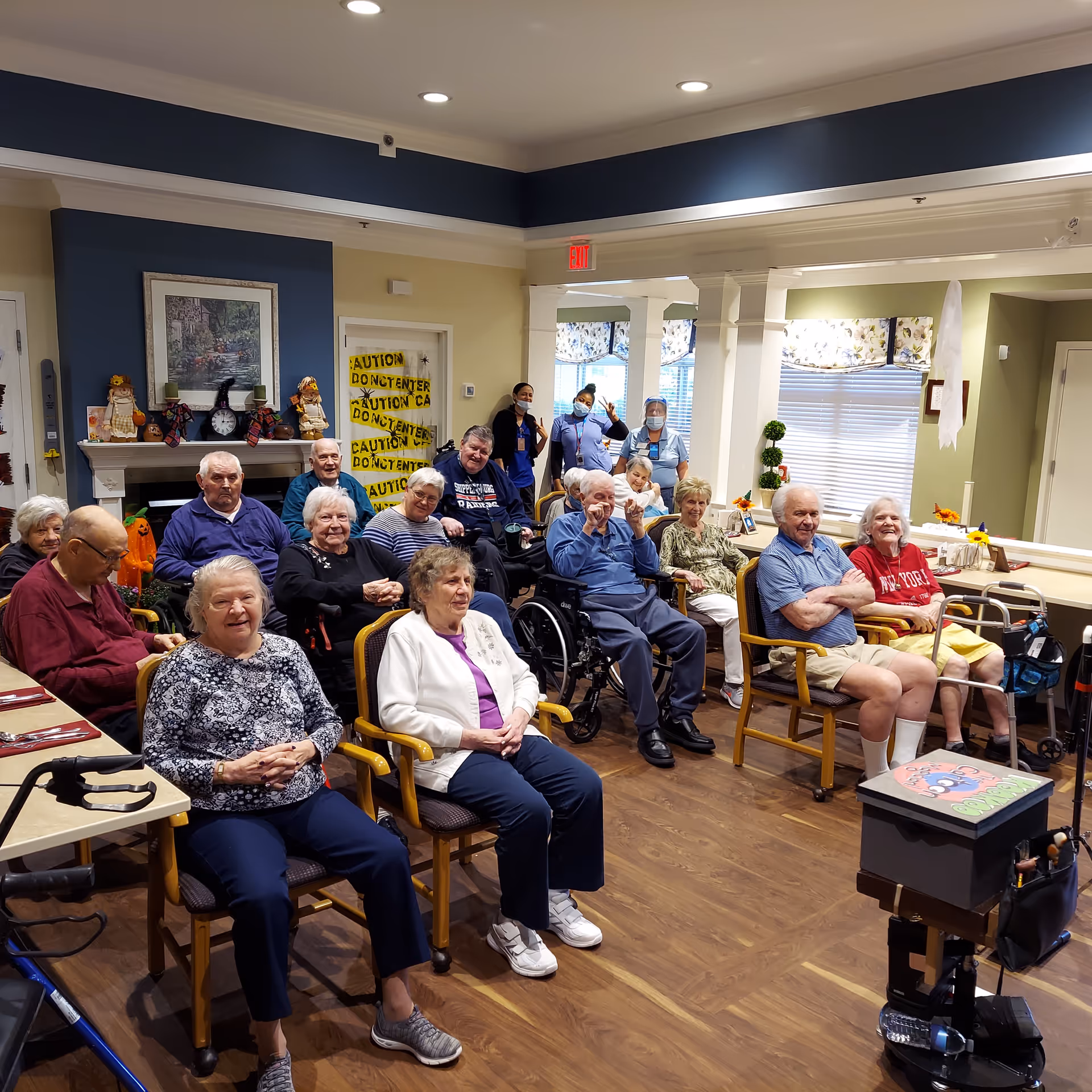 A group of elderly residents seated in chairs and wheelchairs in a common room of a senior living facility. Three staff members wearing masks and face shields stand in the background. The room has wooden flooring, a fireplace decorated with autumn-themed items, and windows with floral curtains. A door in the background is covered with yellow caution tape.
