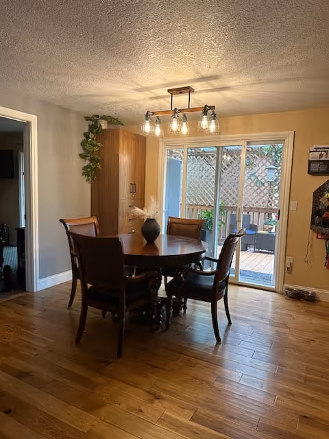 A dining area with a round wooden table and four wooden chairs with cushions. A black vase with dried flowers is placed on the table. There is a modern light fixture with multiple bulbs hanging from the ceiling. In the background, there is a sliding glass door leading to an outdoor patio with lattice fencing and some plants. A tall wooden cabinet with a plant on top is positioned against the wall to the left of the sliding door.