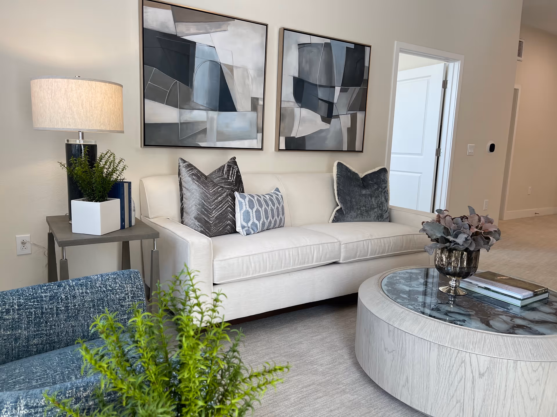 A modern living room with a white sofa adorned with three decorative pillows in shades of gray and blue. Above the sofa are two abstract black, white, and gray paintings. To the left of the sofa is a side table with a lamp, a small plant in a white square pot, and some books. In front of the sofa is a round coffee table with a glass top, holding a decorative plant and a book. A blue textured chair and a green plant are partially visible in the foreground. The room has light-colored walls and carpet, with a doorway visible in the background.