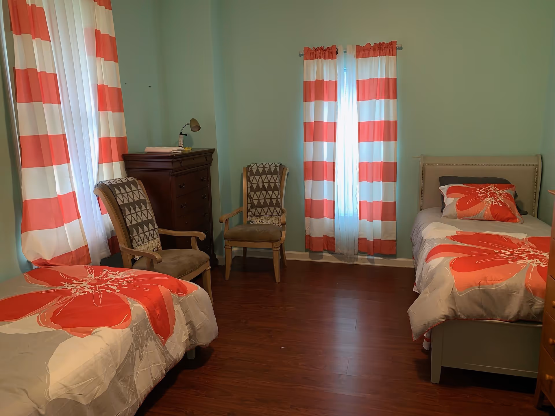A bedroom with two single beds, each with a gray and red floral bedspread. The room has light green walls and wooden flooring. There are two windows with red and white striped curtains. Between the windows, there are two wooden chairs with patterned blankets draped over the backs. A dark wooden chest of drawers with a small lamp on top is positioned near one of the windows.
