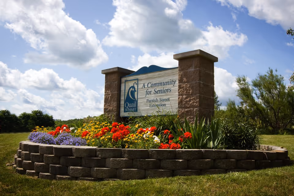 A stone sign for Quail Summit, a community for seniors, surrounded by a flower bed with colorful flowers including red, yellow, and purple blooms, set against a partly cloudy sky and green landscape.