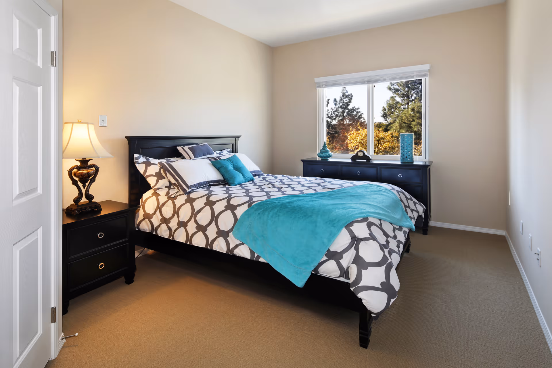 Sunlit bedroom with a queen bed dressed in patterned bedding with teal accents, a bedside table and lamp, and a dresser under a window showing trees outside.