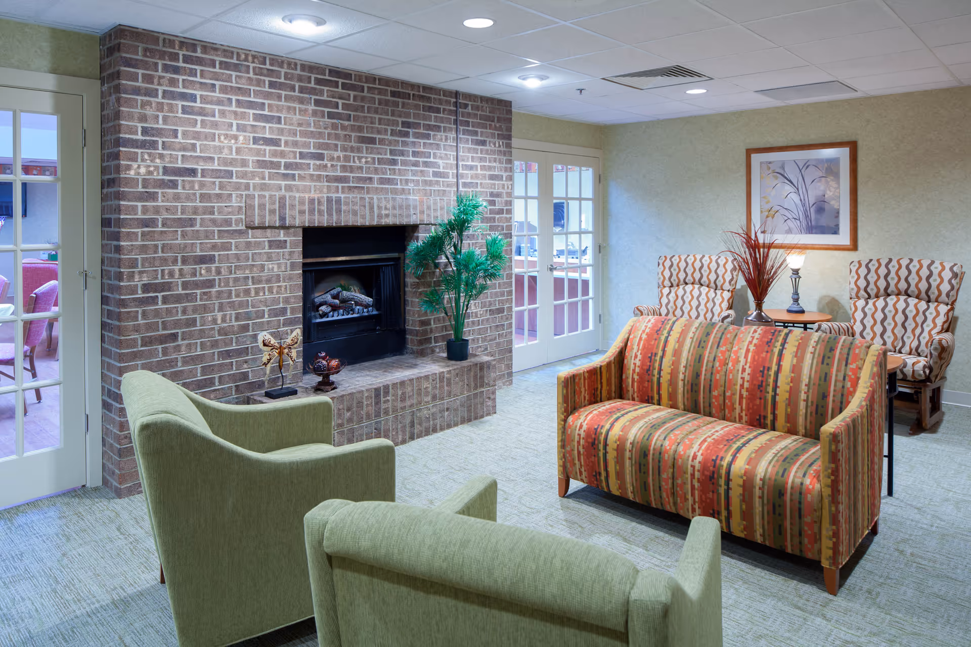 A cozy living room area with a brick fireplace, two green armchairs, a striped sofa, and two patterned armchairs around a small table with a lamp and decorative vase. There are double glass doors leading to another room with pink chairs.
