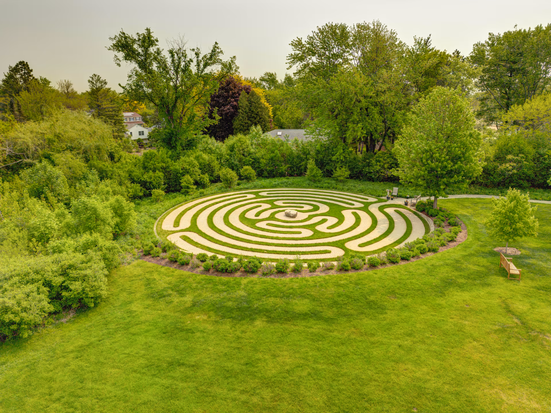 Aerial view of a circular outdoor labyrinth garden surrounded by green grass and trees, with a bench and a walking path nearby.
