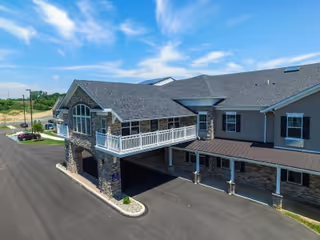 Exterior view of a senior living facility building with stone and siding facade, featuring a covered entrance and a balcony on the second floor under a partly cloudy sky.