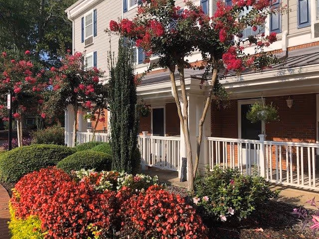 Exterior view of a senior living facility with a white railing porch, brick and beige siding walls, and multiple windows. The foreground features well-maintained landscaping with vibrant red and pink flowers, green shrubs, and small trees with pink blossoms.