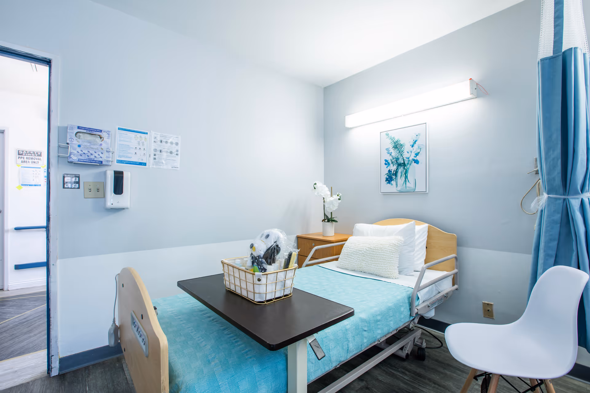 A clean and bright hospital or care facility room with a single bed covered in a light blue blanket and white pillows. A small wooden nightstand with a white orchid plant is next to the bed. Above the bed is a framed picture of blue flowers and a long fluorescent light fixture. A black overbed table holds a basket with various items. A white chair is positioned near the bed, and blue privacy curtains hang to the right. The door to the room is open, showing a hallway with signs on the wall.