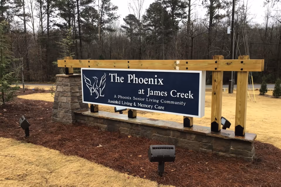 Outdoor sign for The Phoenix at James Creek, a senior living community offering assisted living and memory care, mounted on a stone and wooden structure with trees and a road in the background.