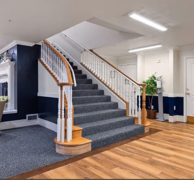 Interior view of a senior living facility showing a carpeted staircase with wooden handrails and white balusters. The area has a mix of carpet and wood flooring, a potted plant near a white door, and a small stand with a dispenser. The walls are painted white with a dark blue lower section, and there is a fireplace partially visible on the left side.