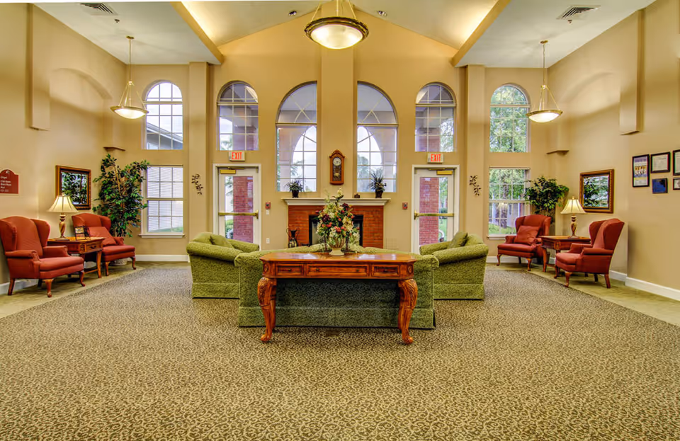 A spacious and well-lit living room area with high ceilings and large arched windows. The room features green upholstered sofas arranged around a wooden coffee table with a floral centerpiece. There are red armchairs with side tables and lamps on either side of the room. The walls are painted beige, and there are plants and framed pictures decorating the space. Two exit doors are visible at the back of the room.