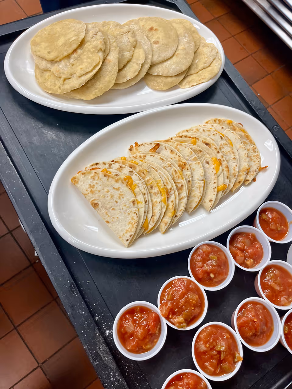 Two white oval plates on a black tray, one plate with folded quesadillas filled with melted cheese, the other plate with small round flatbreads. Several small white cups filled with chunky red salsa are arranged next to the plates.