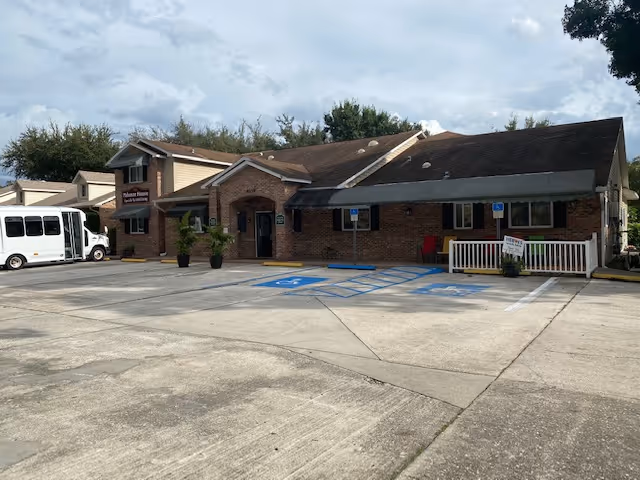 Exterior view of Palamar House, a single-story brick building with a sloped roof and an entrance covered by a small awning. There are two handicap parking spaces in front, a white bus parked on the left side, and some potted plants near the entrance. The sky is partly cloudy.