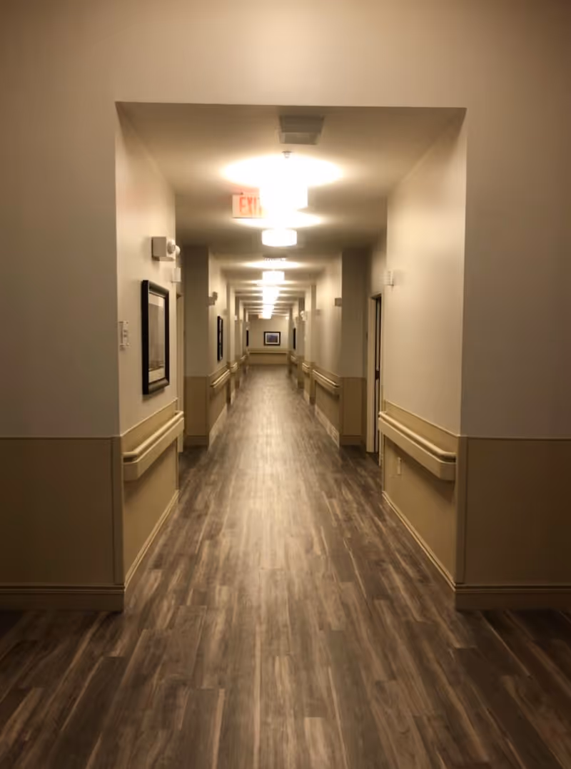 A long, well-lit hallway in a senior living facility with wood-patterned flooring, beige walls with handrails on both sides, framed pictures hanging on the walls, and multiple ceiling lights leading to a distant end.