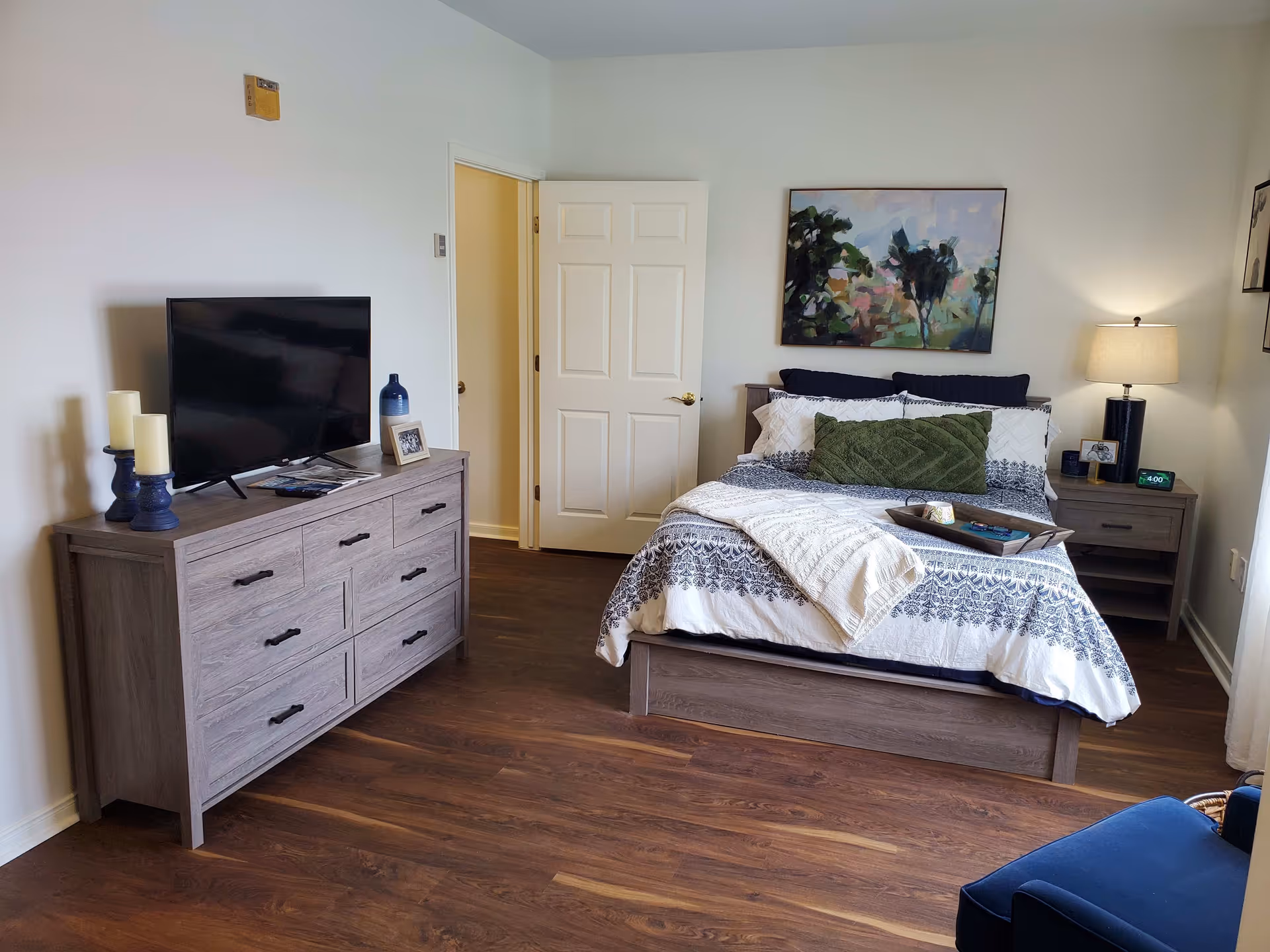 A furnished bedroom with a bed, nightstand and lamp, dresser with a TV, and wall art on hardwood floors.