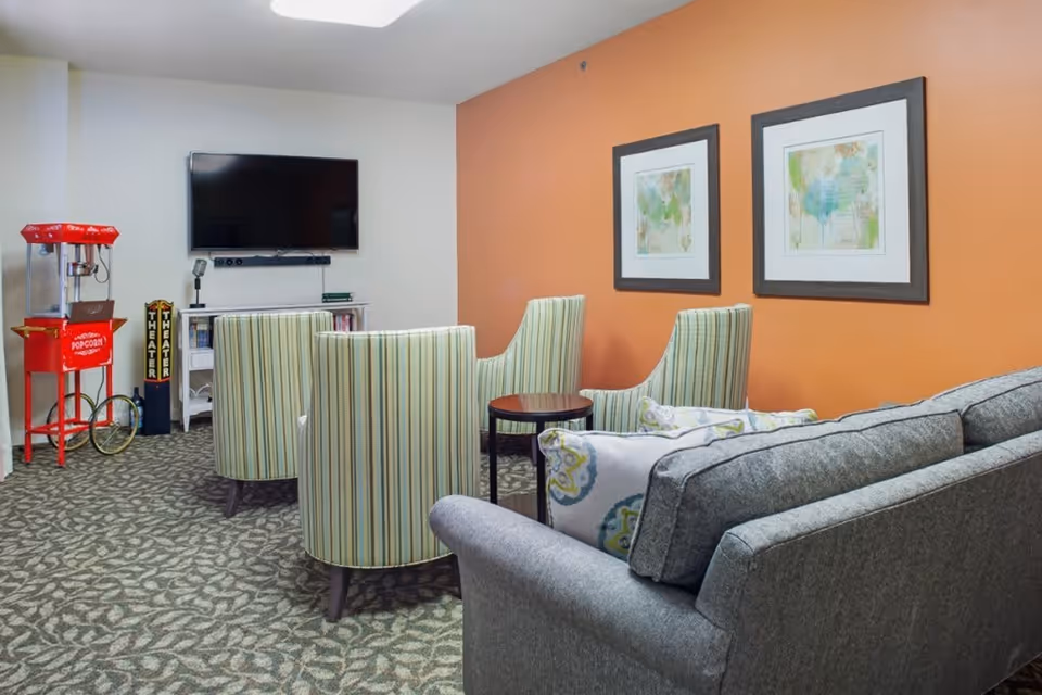 A cozy living room area with a gray couch and three striped armchairs arranged around a small round table. A flat-screen TV is mounted on the wall above a white media console. There is a red popcorn machine on wheels in the corner and two framed abstract paintings on an orange accent wall.