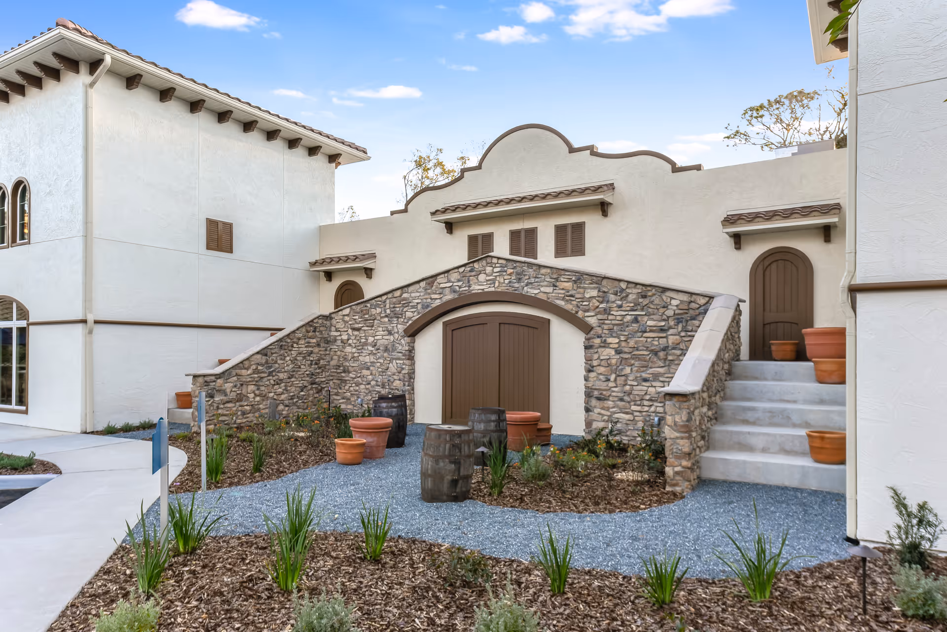 Exterior view of Tuscan Gardens of Palm Coast Senior Living showing a building with beige stucco walls, stone accents, and brown wooden doors. There are stairs leading up to a door on the right side, several terracotta pots, and landscaped garden beds with small plants and mulch. The sky is blue with a few clouds.