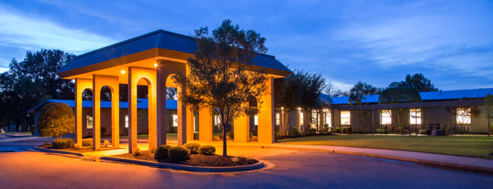 Exterior view of Stella Manor facility at dusk with illuminated entrance featuring tall brick pillars and a covered driveway, surrounded by trees and landscaped bushes.