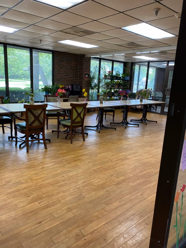 A bright room with large windows showing greenery outside. Inside, there are several tables arranged in a row with chairs around them. Each table has a small vase with colorful flowers. There are plants on shelves near the windows and a wooden floor with a light finish.