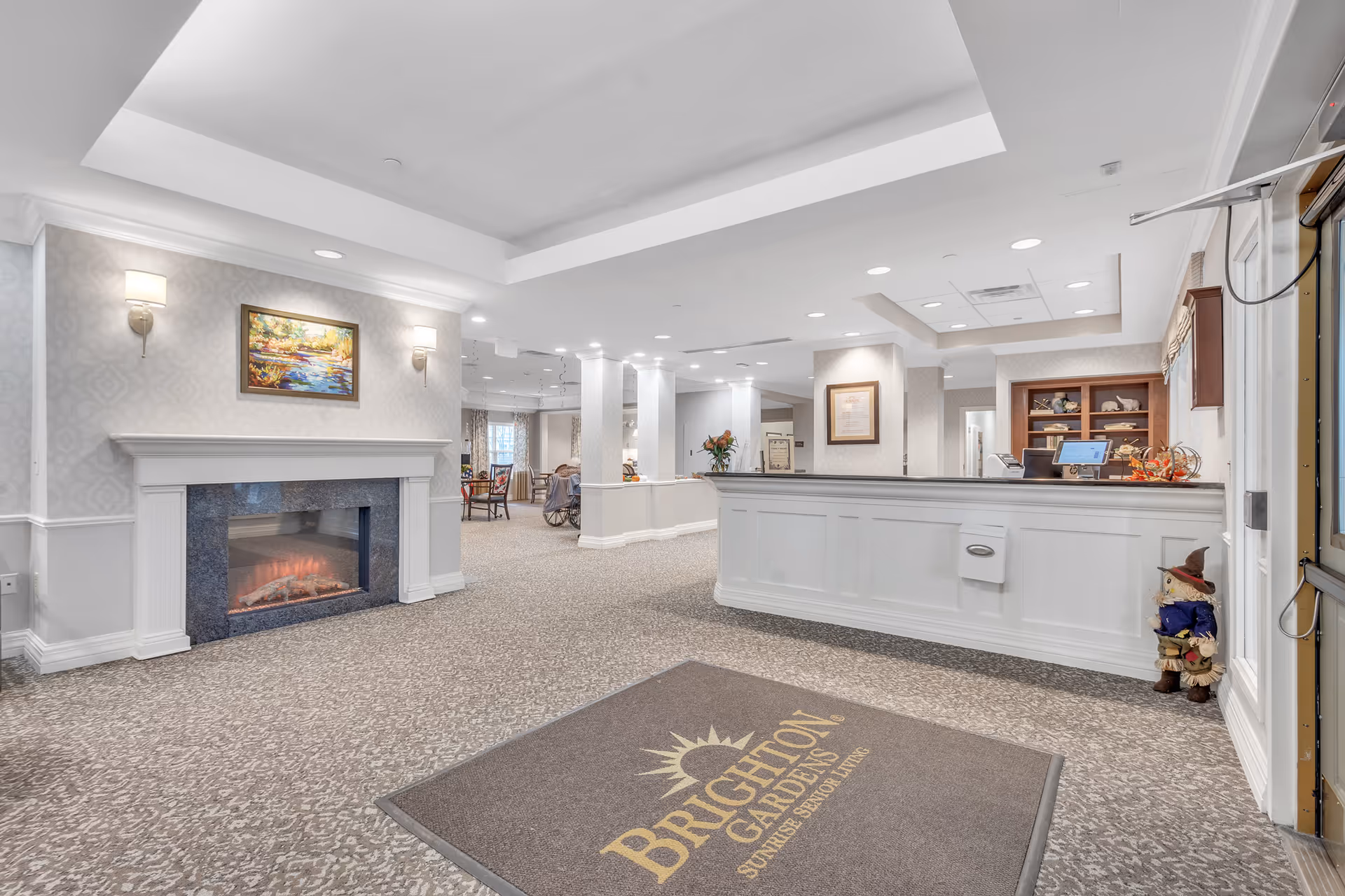 Reception area of Brighton Gardens of Florham Park featuring a white front desk, a fireplace with a painting above it, wall sconces, and a carpet with the facility's logo. The space is well-lit with recessed lighting and decorated with flowers and a small scarecrow figure near the entrance.