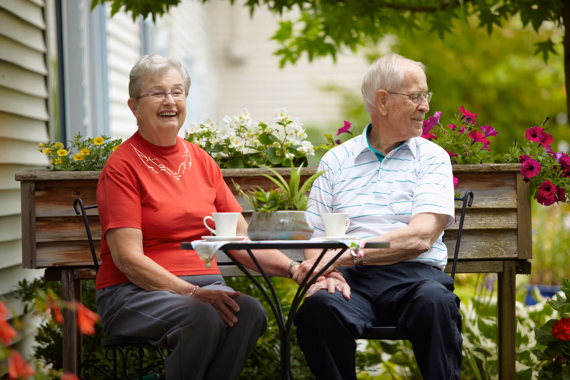 An elderly couple sitting outdoors at a small round table with two white cups. The woman, wearing a red shirt and glasses, is smiling and looking forward. The man, wearing a white striped polo shirt and glasses, is looking to the side. Behind them is a wooden planter box filled with colorful flowers and greenery.