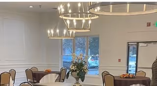 A dining room with round tables covered in white and brown tablecloths, each table surrounded by chairs. A large floral centerpiece is on one table. Modern chandeliers hang from the ceiling. Windows and a door are visible in the background.