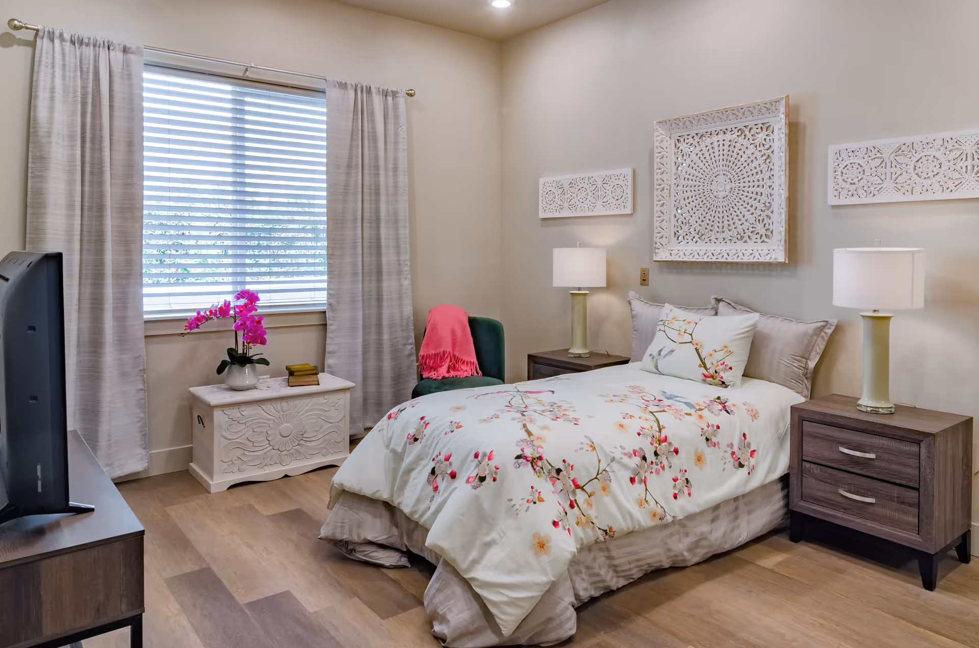 A cozy bedroom with a single bed featuring floral bedding and decorative pillows. The room has two bedside tables with lamps, a green chair with a pink throw, a white chest with a pink orchid and books on top, and a window with white blinds and light-colored curtains. The walls are decorated with three white carved wooden panels.