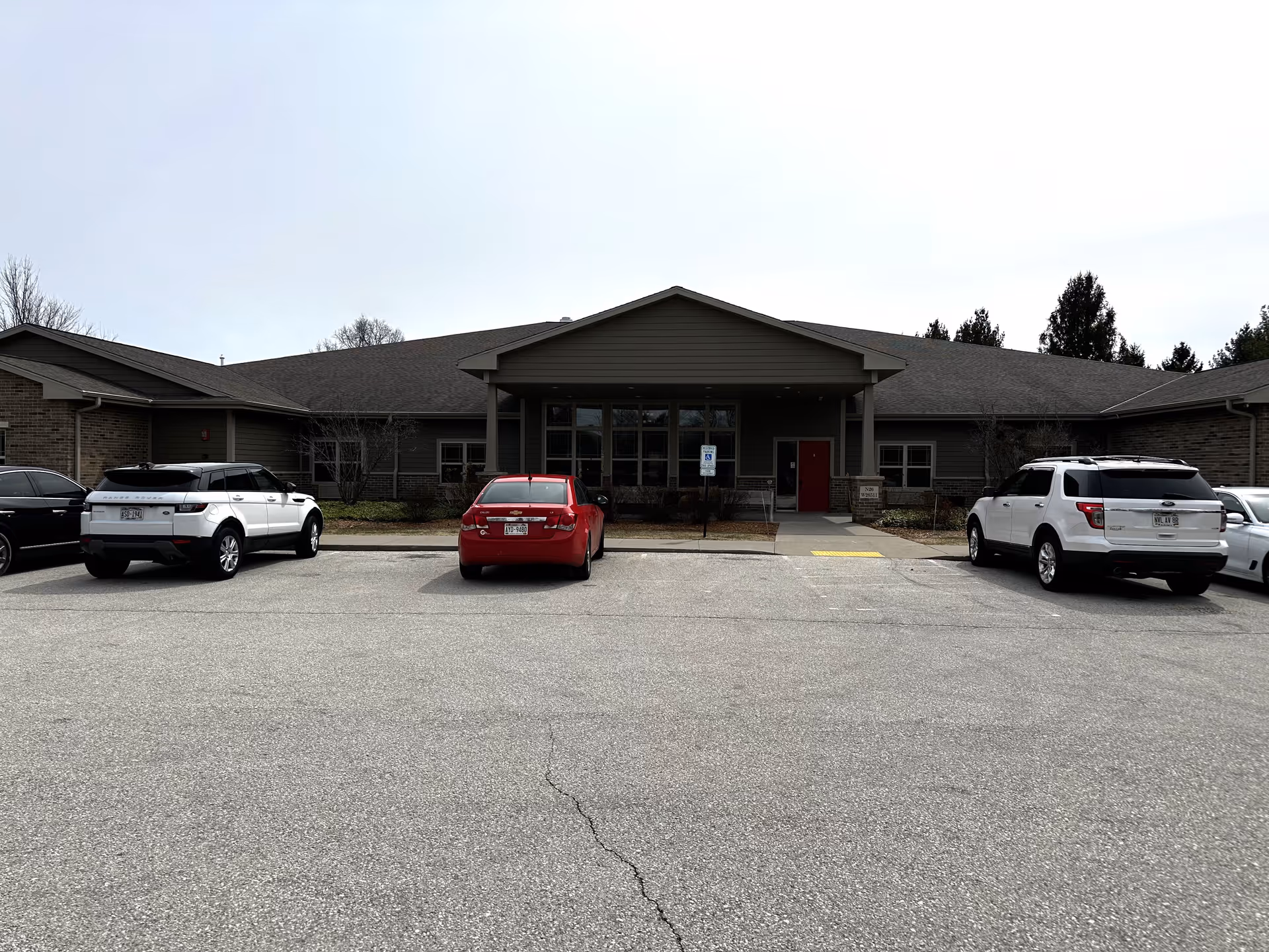 Front exterior view of a single-story building with a covered entrance and several parked cars in front. The building has a gray roof and siding, with a red door visible near the entrance. Trees and shrubs surround the building.