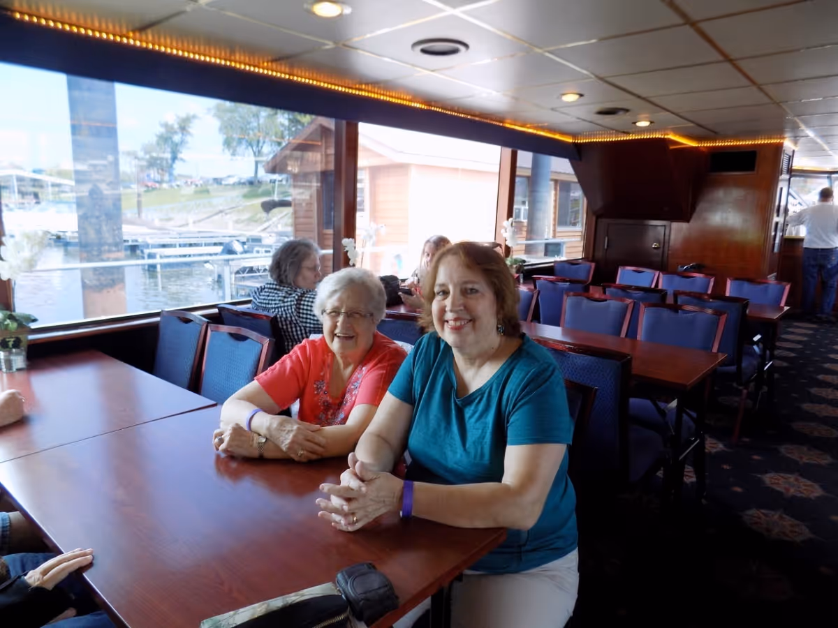 Two women sitting at a wooden table inside a dining area with large windows showing a waterfront view outside. The room has multiple tables and chairs, and other people are seated in the background.