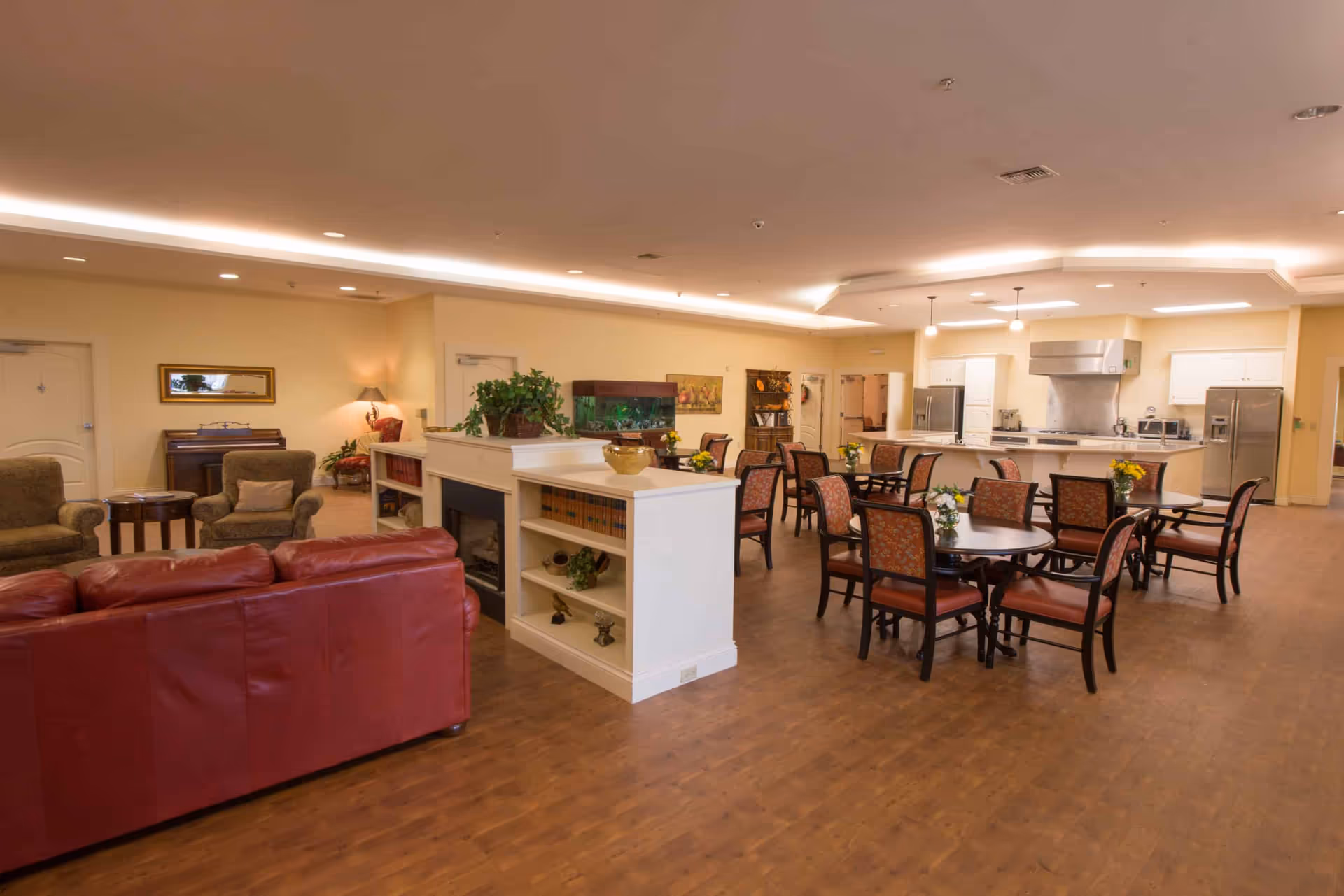Communal dining and lounge area with multiple round dining tables and chairs, a kitchen in the background, and a red sofa in the foreground.
