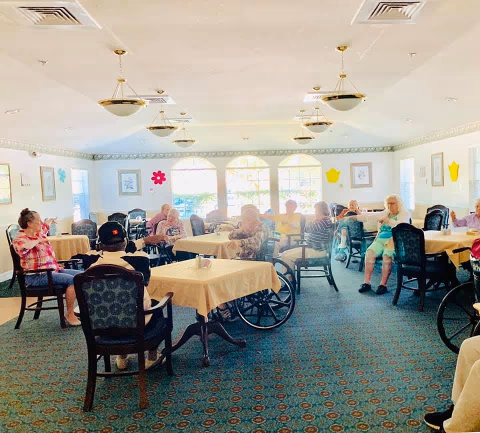 A group of elderly people seated around tables in a bright, spacious room with large windows. The room has patterned carpet, beige tablecloths on the tables, and decorative wall hangings. Some individuals are in wheelchairs, and they appear to be engaging in a group activity or exercise.