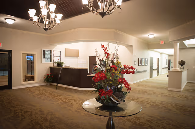 Interior view of a senior living facility lobby area with a round glass table holding a large floral arrangement with red and green flowers. There is a reception desk in the background with a plant on it, beige walls, carpeted floor, and ceiling lights including chandeliers. A hallway extends to the right with framed pictures on the walls.