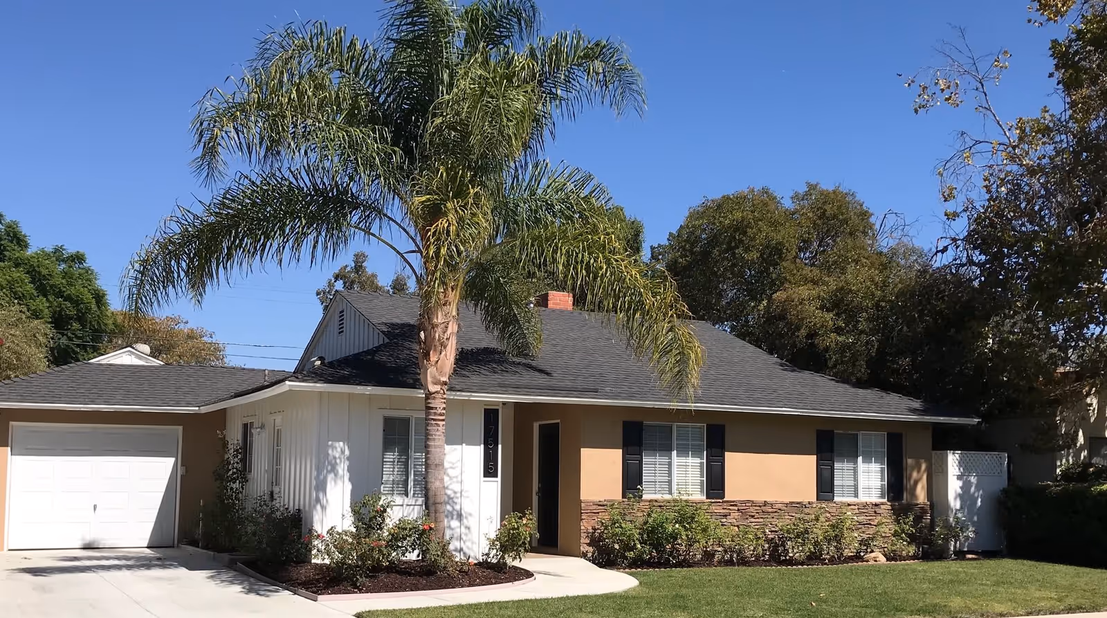 Single-story residential building with a light brown exterior, white garage door, black shutters, and a black roof. A tall palm tree and various shrubs are planted in front of the house under a clear blue sky.