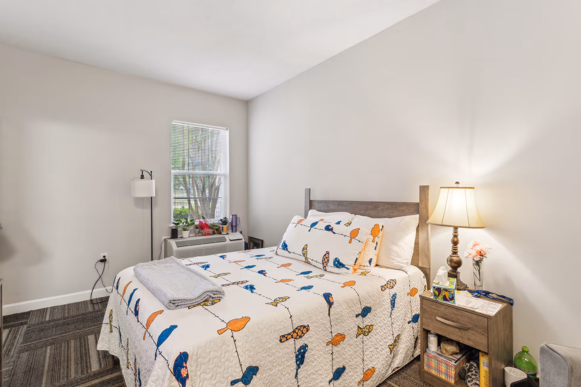 Bright bedroom featuring a bed with a colorful bird-pattern quilt, wooden headboard, a bedside table with lamp, a window and floor lamp.