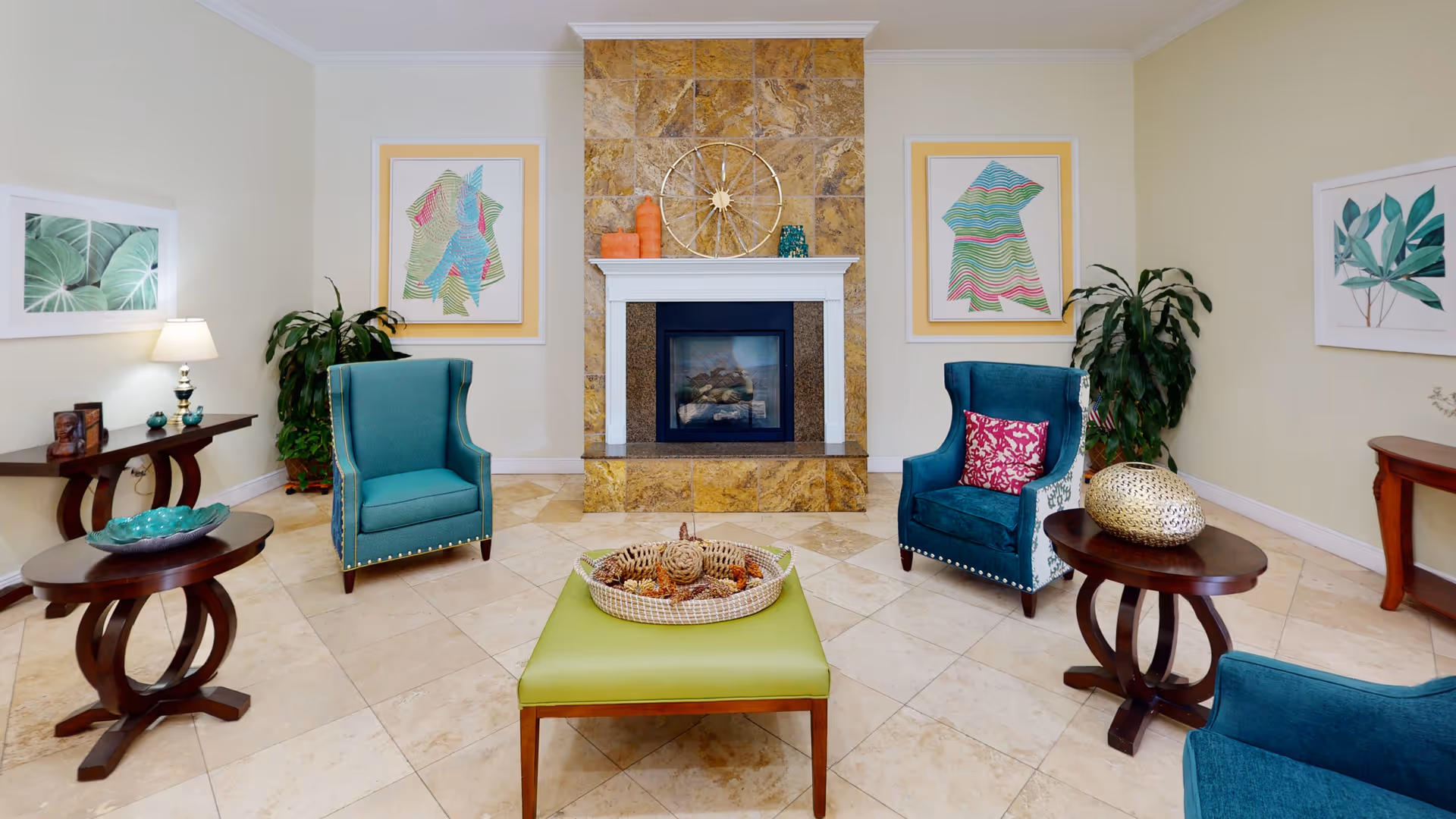Bright communal living room with teal armchairs arranged around a green ottoman and a central fireplace, decorated with side tables, plants, and wall art.