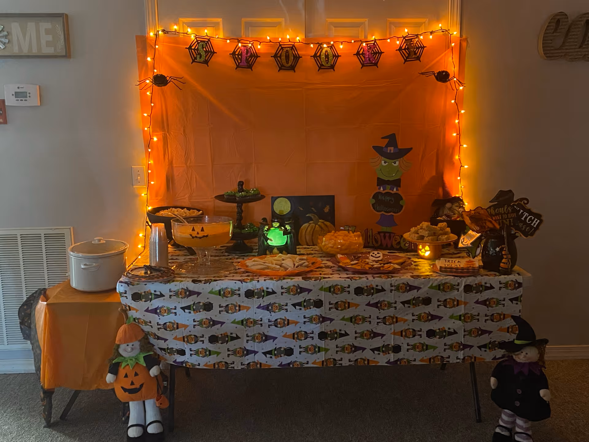 A Halloween-themed table setup with orange string lights and a backdrop decorated with spider webs and spiders. The table is covered with a Halloween patterned tablecloth and holds various snacks, a punch bowl with a jack-o'-lantern face, and Halloween decorations including a green glowing jar, a pumpkin, and a witch figure. Two doll-like figures dressed in Halloween costumes stand on either side of the table.