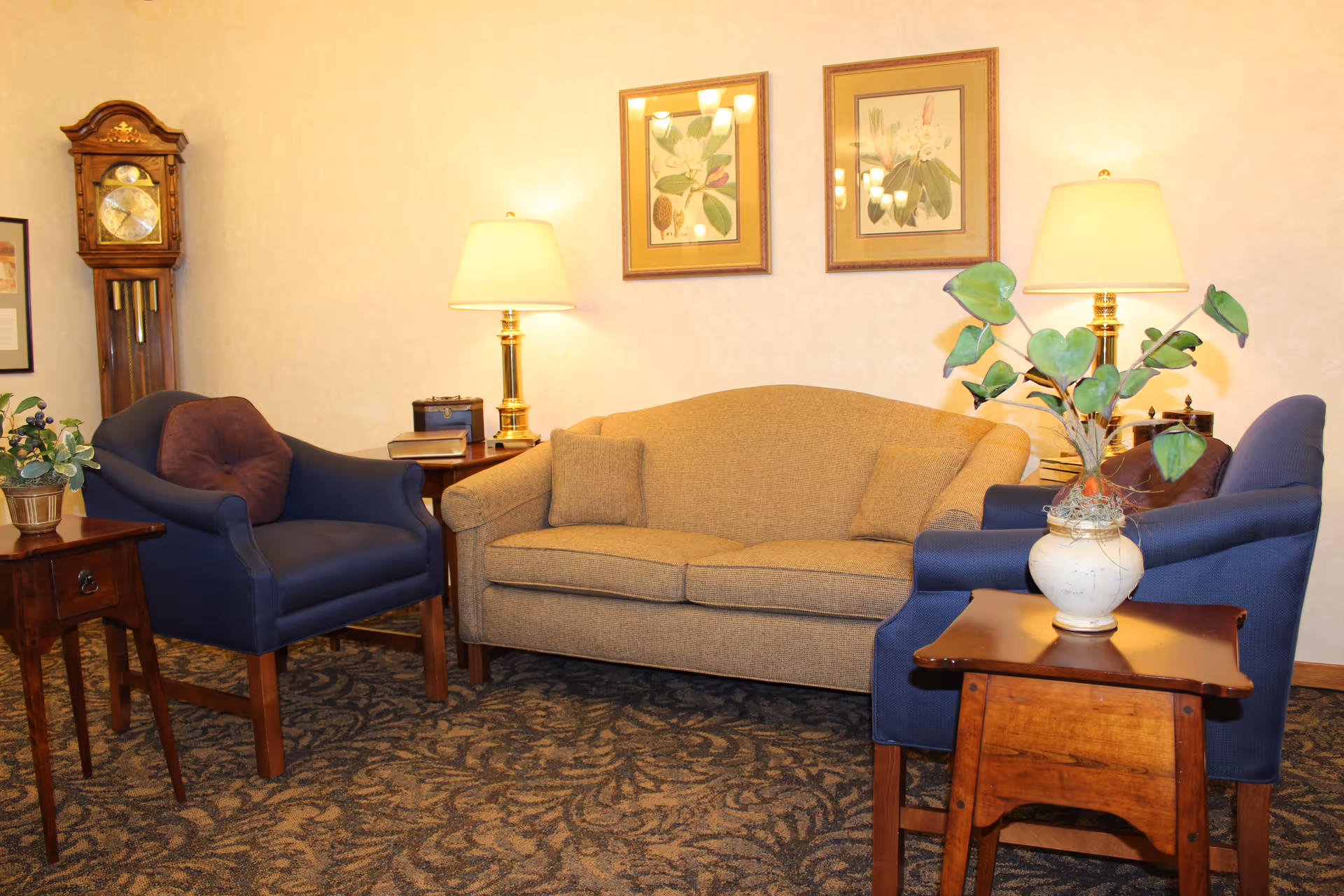 A cozy living room area with a beige sofa flanked by two blue armchairs, each with a brown cushion. There are two wooden side tables with table lamps on either side of the sofa, a small wooden table with a potted plant in the foreground, and a grandfather clock in the corner. Two framed botanical prints hang on the wall above the sofa.