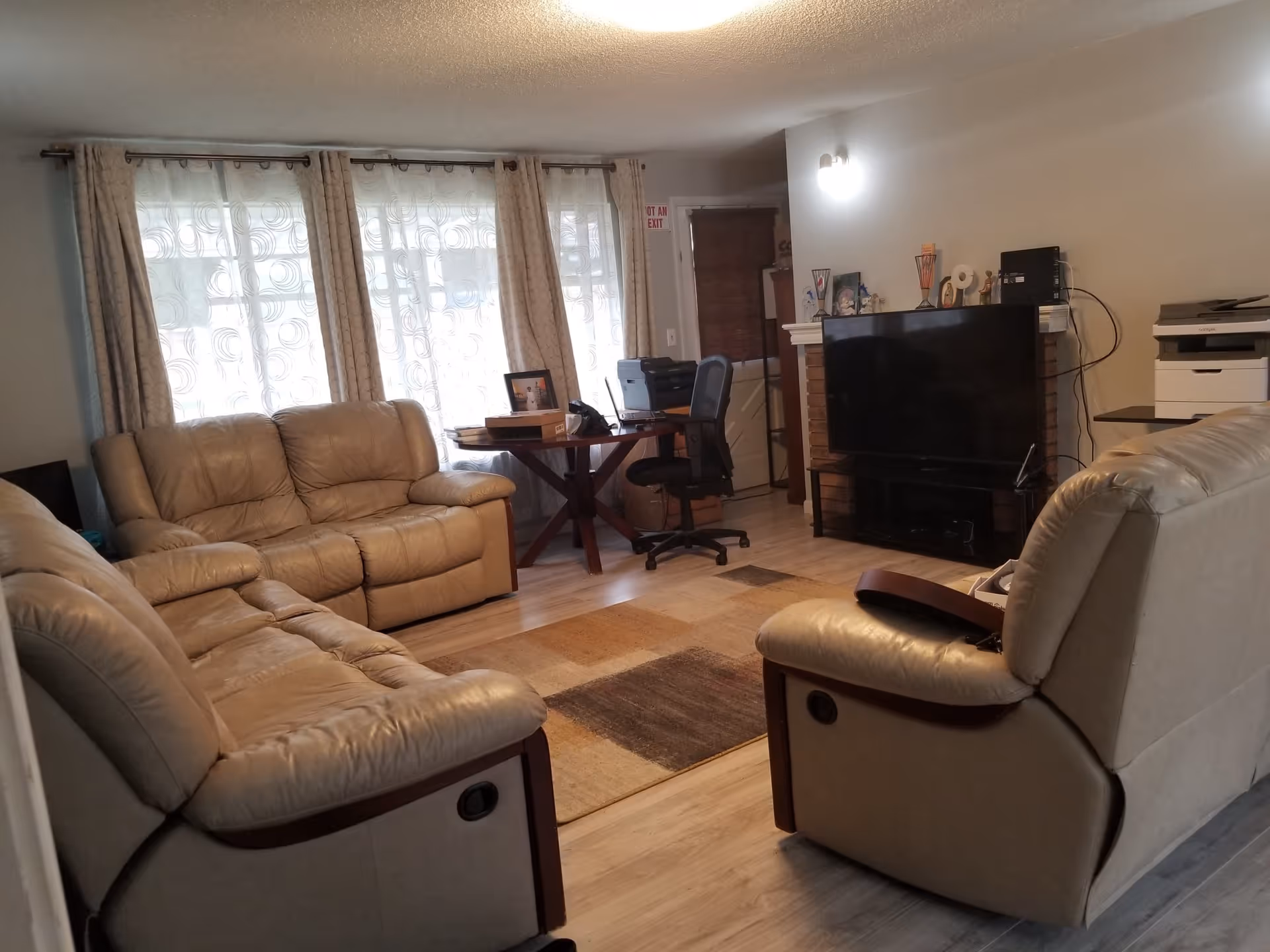 A living room with beige leather sofas arranged around a rectangular area rug. There is a large flat-screen TV on a stand in front of a brick fireplace. A wooden table with a black office chair and a printer is positioned near a window with sheer and patterned curtains. The room has light-colored wooden flooring and neutral-colored walls.