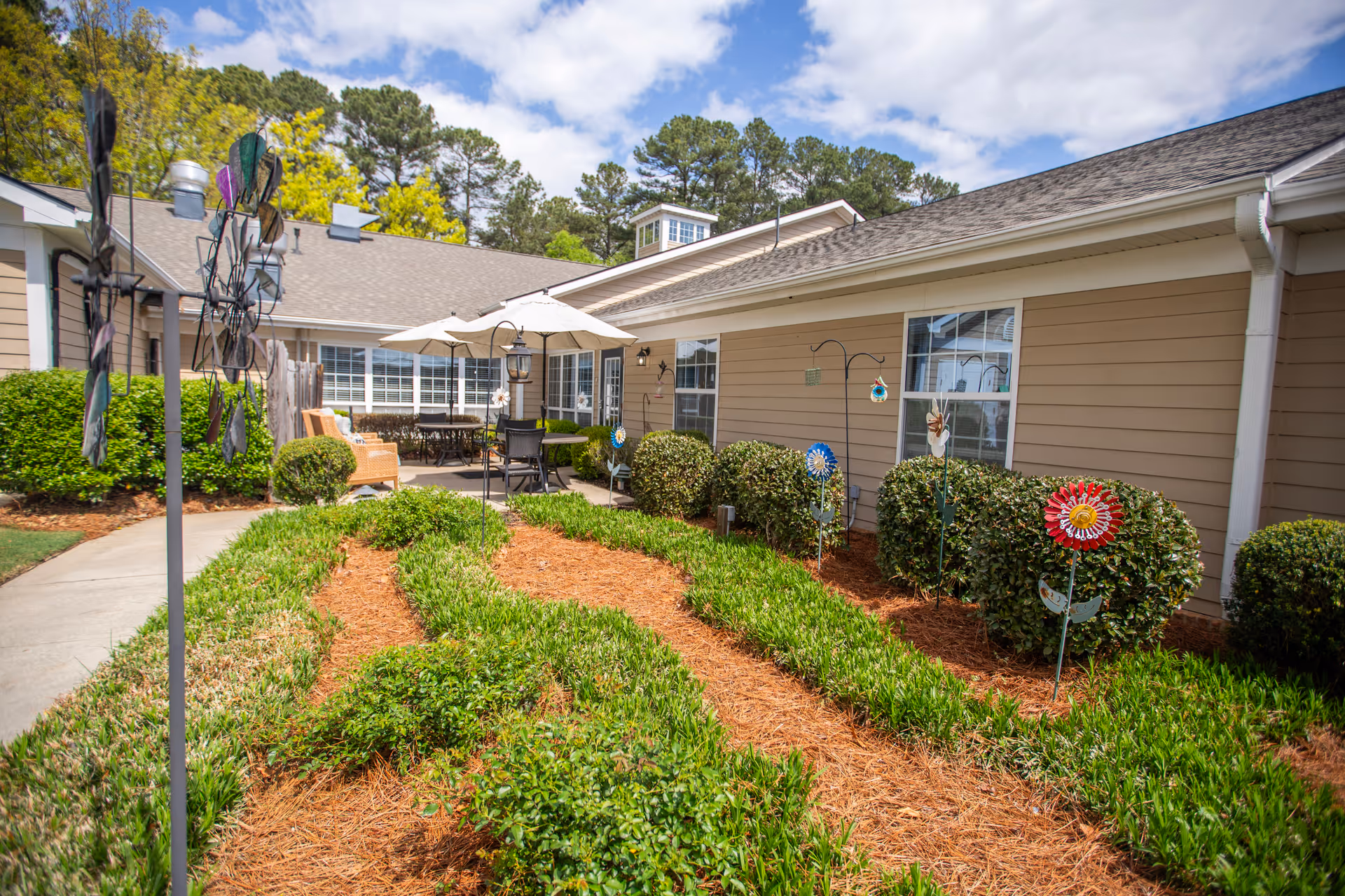Outdoor garden area at a senior living facility with green shrubs, decorative garden stakes, and a patio with tables, chairs, and umbrellas under a partly cloudy sky.