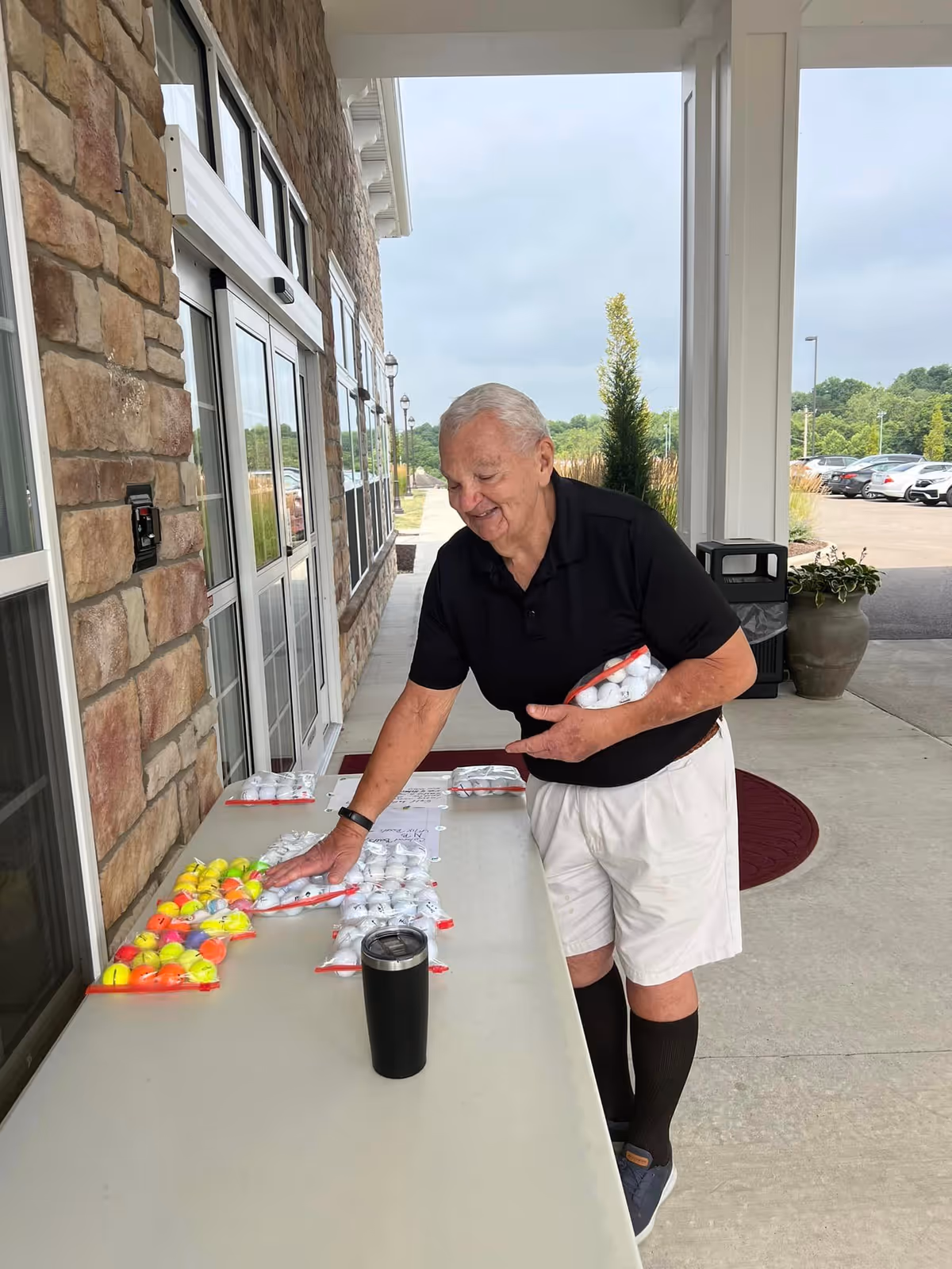 An elderly man wearing a black polo shirt, white shorts, and black knee-high socks is standing outside a building with stone walls. He is smiling and organizing multiple bags of golf balls on a table. There is a black tumbler on the table and a parking lot with cars in the background under a cloudy sky.