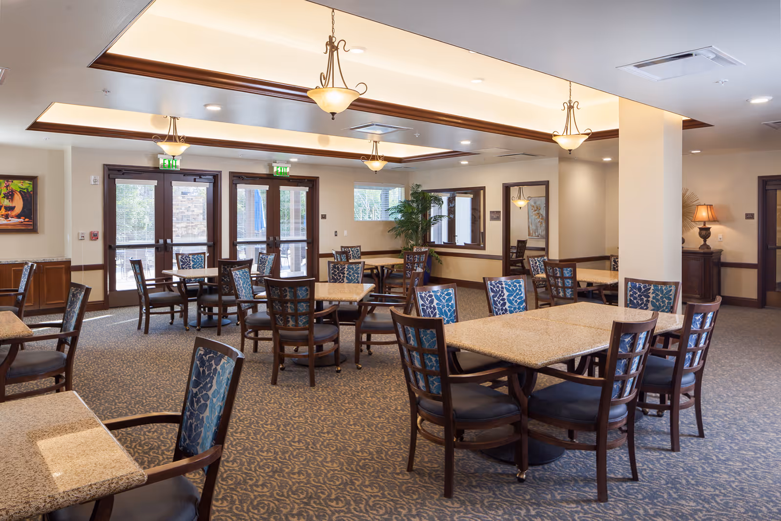 A spacious dining room in a senior living facility with multiple tables and chairs arranged neatly. The room features carpeted floors, warm lighting from ceiling fixtures, large windows, and glass doors leading outside. There are decorative plants and framed artwork on the walls, creating a welcoming atmosphere.
