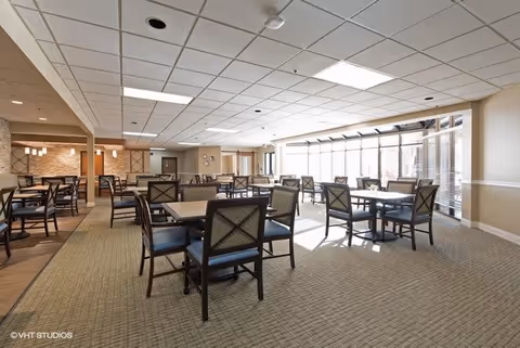 A spacious dining room with multiple tables and chairs arranged neatly. The room has large windows on one side allowing natural light to fill the space. The ceiling has recessed lighting and a grid pattern. The floor is carpeted, and the walls are painted in neutral tones.